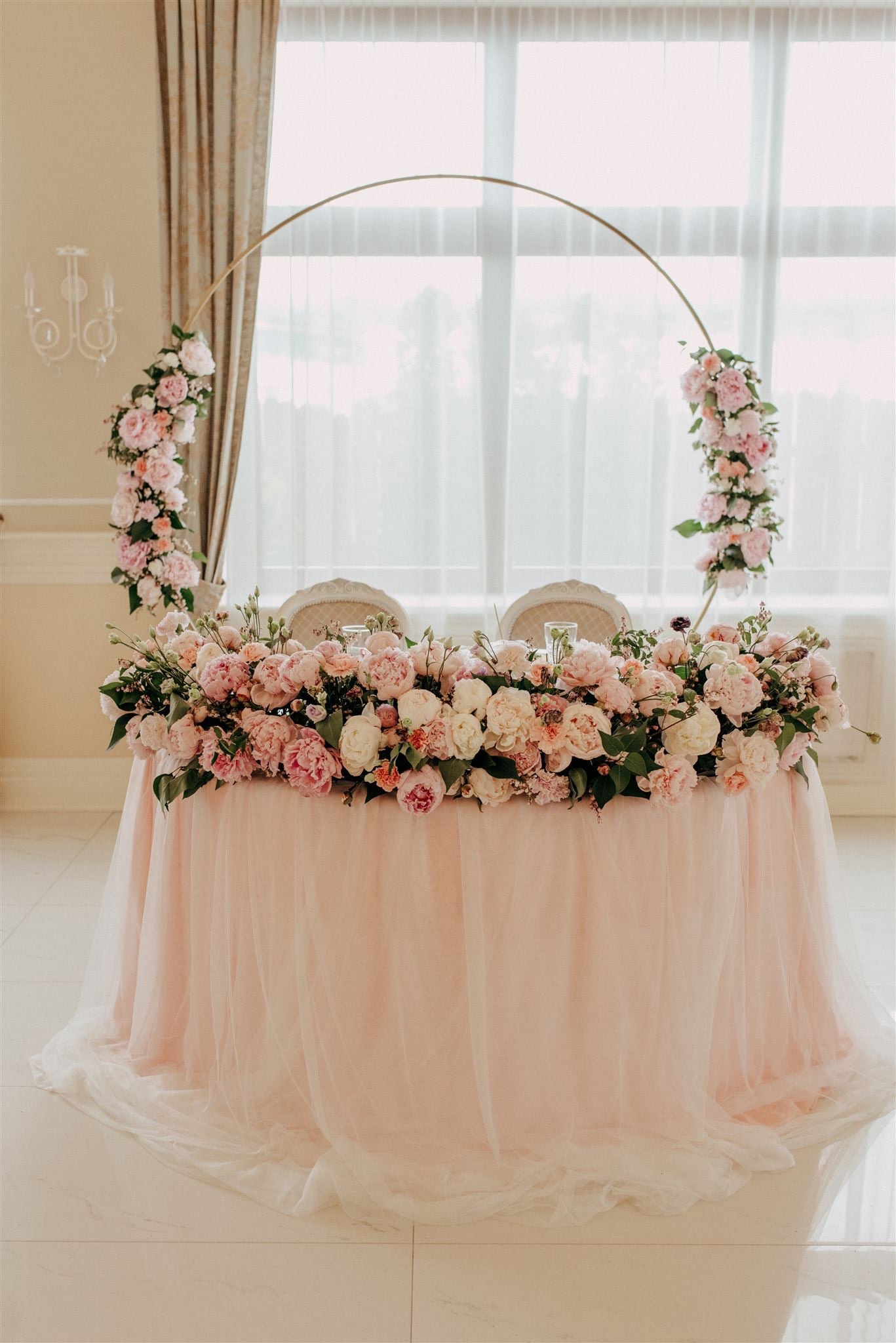 Stół weselny udekorowany pastelowymi różami i zielenią, z okrągłym złotym stelażem również ozdobionym kwiatami, ustawiony na tle okna z białymi zasłonami.