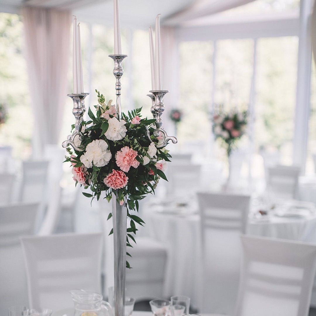 Elegancki, srebrny świecznik z kompozycją z białych i różowych goździków na stole w sali bankietowej z białymi krzesłami i oknami w tle.