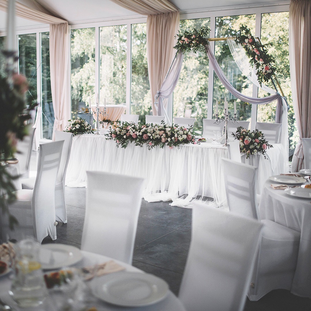 Uroczysta sala z długim stołem prezydialnym udekorowanym białym obrusem z falbanami, kompozycjami kwiatowymi z różowych i białych róż, srebrnymi świecznikami; tło stanowią okna z widokiem na zieleń...