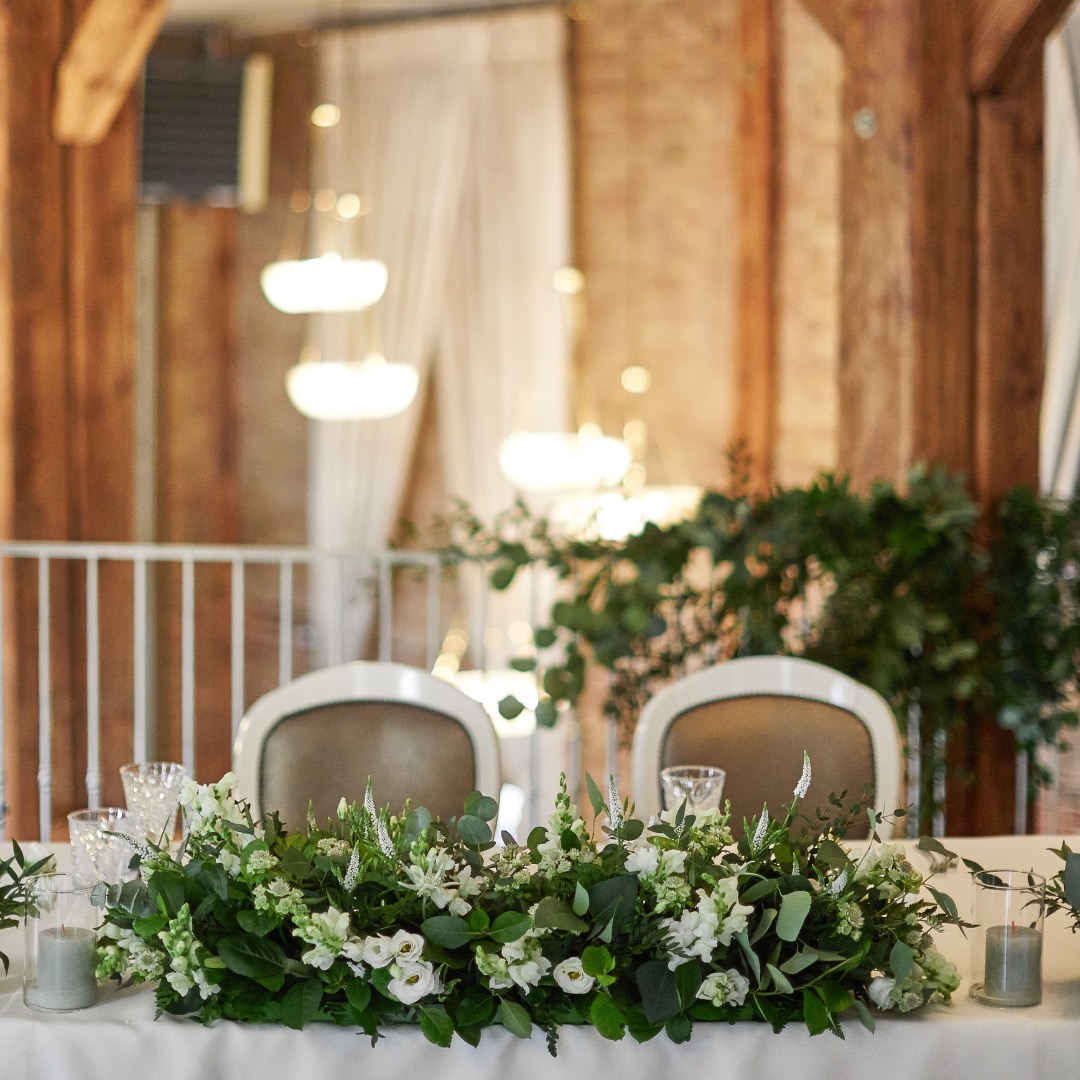Dekoracja stołu weselnego z białymi kwiatami i zielonymi liśćmi, szklane naczynia i świece, w tle drewniana konstrukcja i białe balustrady.
