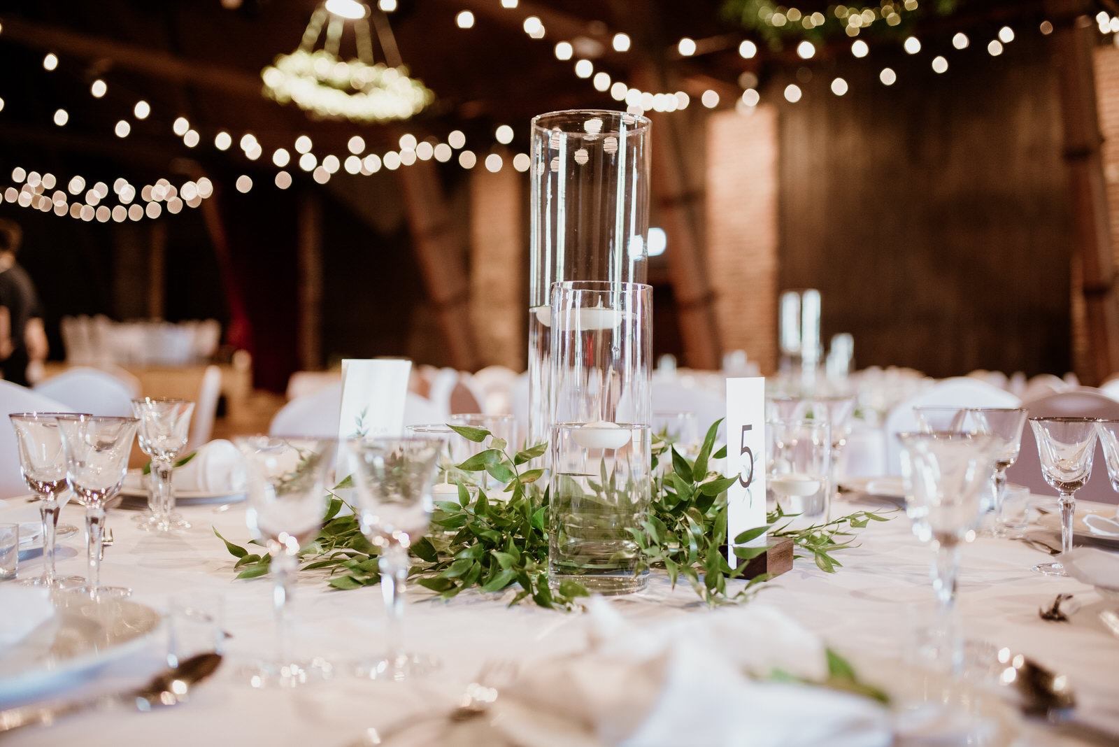Uroczysty stół z numerem '5', ozdobiony wysokimi szklanymi wazonami z pływającymi świecami, otoczony zielonymi liśćmi, nakryty białym obrusem i kryształowymi kieliszkami, w tle girlanda z żarówek.