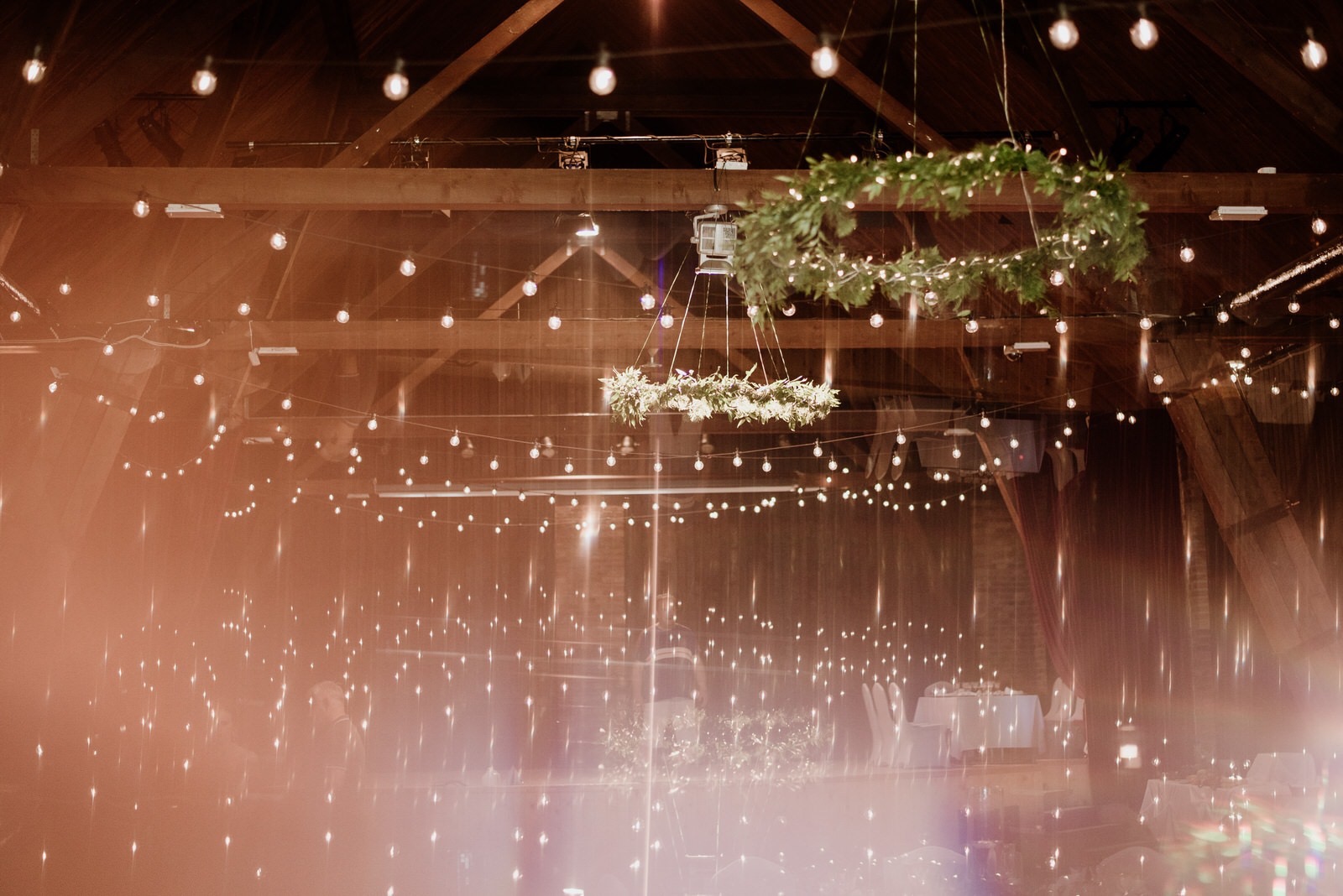 Dekoracja sali weselnej z drewnianą konstrukcją, girlandami z żarówek i wiszącymi wiankami z zieleni i kwiatów, widok z dołu z efektem rozmycia.