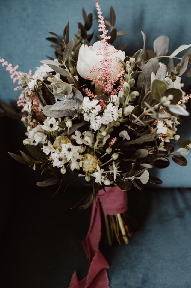 Delikatny bukiet ślubny z białych peonii, drobnych białych kwiatów i różowych akcentów, przewiązany bordową wstążką na tle turkusowej tkaniny.