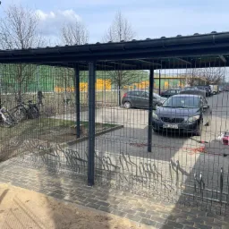 Zadaszona wiata rowerowa z metalową konstrukcją i kostką brukową, widok z zewnątrz na zaparkowane rowery i samochody, w tle budynek z zielono-żółtą elewacją.