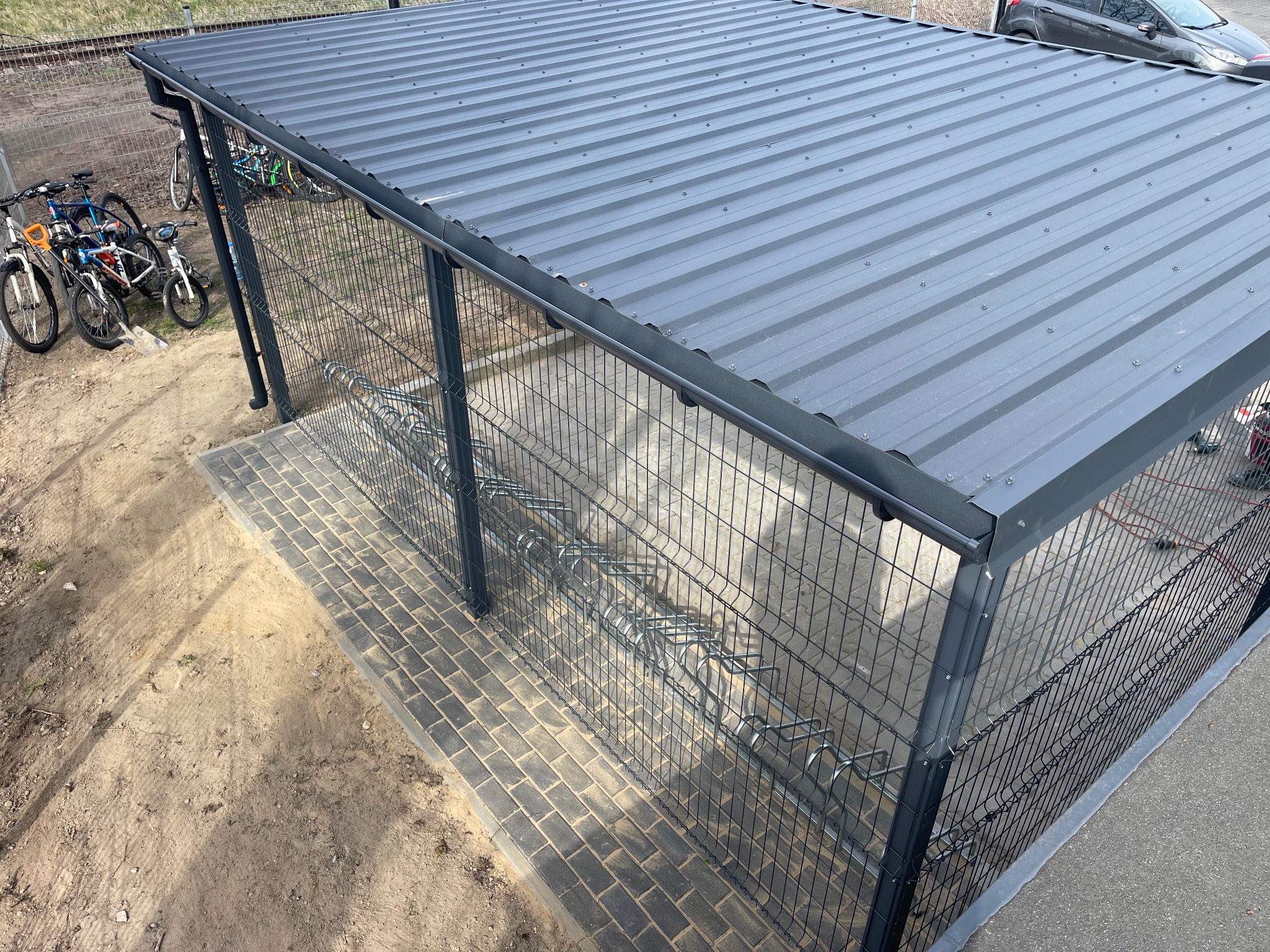 Zadaszona wiata rowerowa z szarą, metalową konstrukcją i siatką, wyłożona kostką brukową, obok zaparkowane rowery i samochód.
