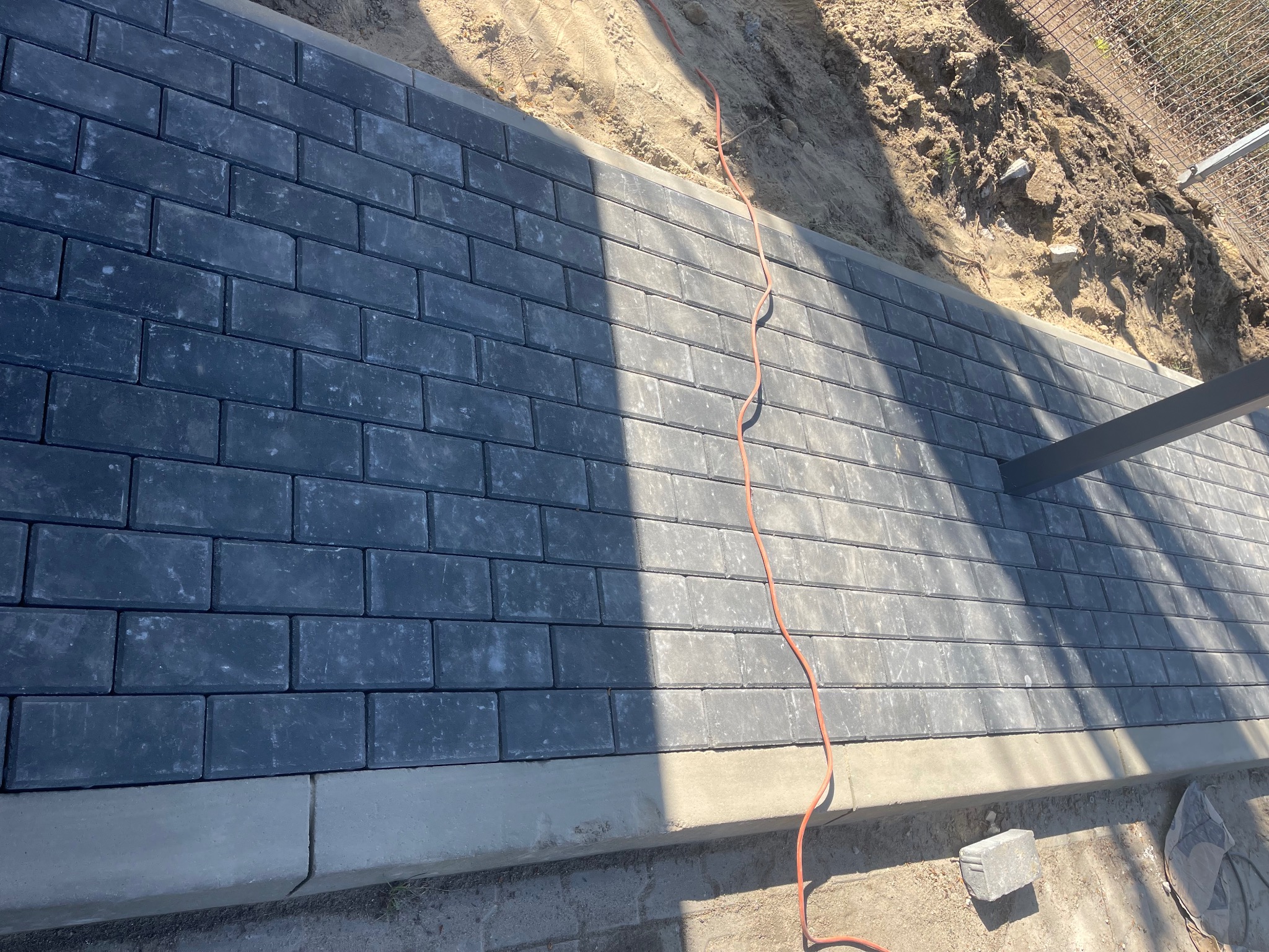 Szara kostka brukowa ułożona w rzędach, obramowana betonowym krawężnikiem, z czerwoną linką elektryczną biegnącą po powierzchni. Widoczny fragment metalowej konstrukcji i ziemia w tle.