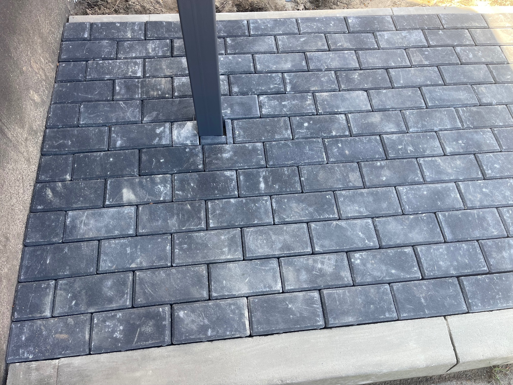 Szara, prostokątna kostka brukowa ułożona wokół metalowego słupka, obrzeże z jasnoszarego betonu.