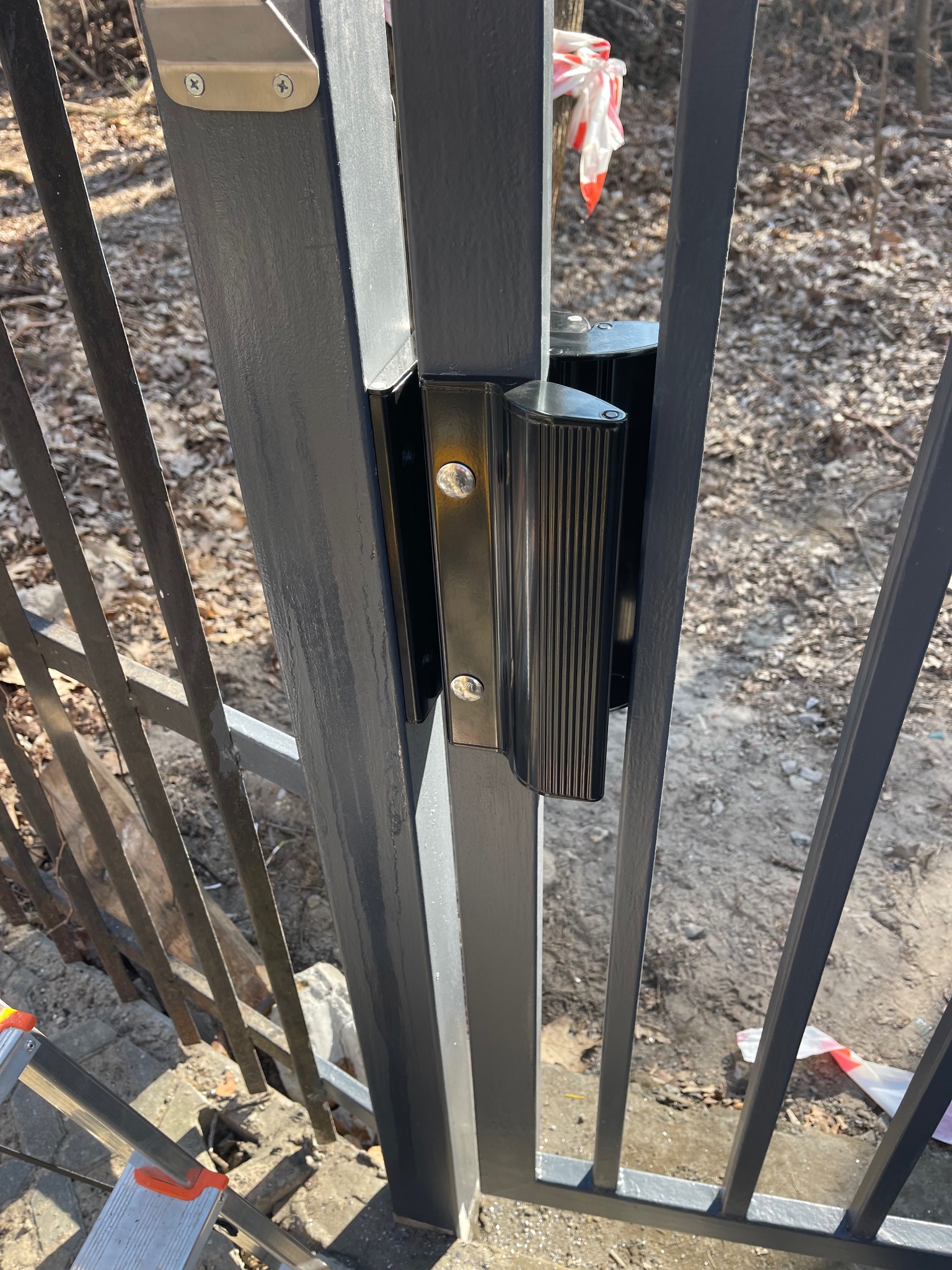 Szczegółowe ujęcie nowo zamontowanej, grafitowej bramy z ryflowaną klamką, widoczne ślady po malowaniu, w tle ziemia i taśma ostrzegawcza.