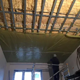Prace wykończeniowe na poddaszu: widoczna izolacja z wełny mineralnej między metalowymi profilami sufitowymi, zabezpieczona folią, pracownik na rusztowaniu. Częściowo widoczne okno i ściany...