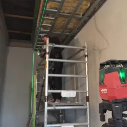 Pracownik na rusztowaniu, używający lasera krzyżowego Hilti PM 40-MG do wyznaczania poziomów przy montażu metalowej konstrukcji sufitu podwieszanego z ociepleniem z wełny mineralnej.