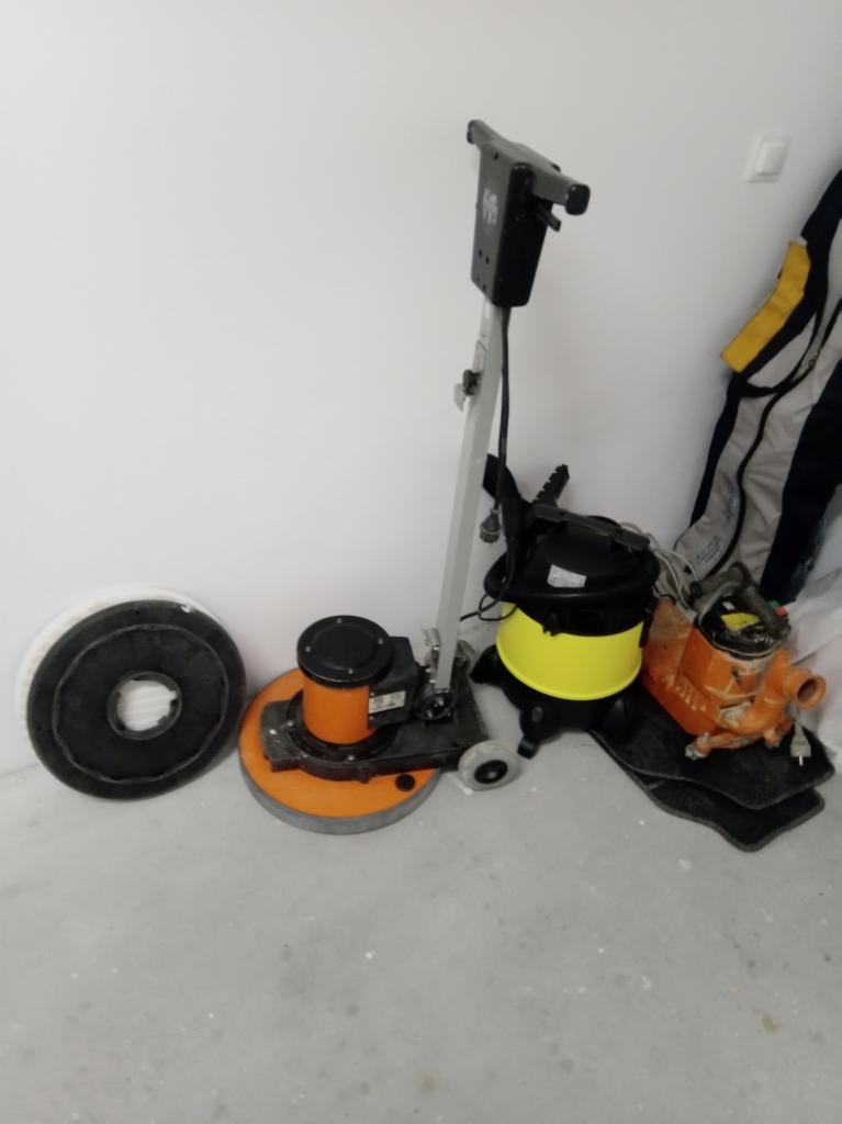 Sprzęt do renowacji podłóg: szlifierka do parkietu z tarczą szlifierską obok, odkurzacz przemysłowy z żółtym zbiornikiem oraz dodatkowe urządzenie do obróbki drewna, wszystko ustawione przy białej...