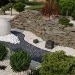 Nowoczesny ogród z białym żwirem, kamienną ścianką, kulistą rzeźbą i ścieżką z czarnego grysu.