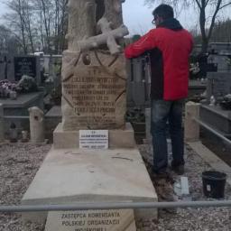 Kamieniarstwo Artystyczne - Pracownik w czerwonej kurtce czyści jasny, kamienny nagrobek z inskrypcjami na cmentarzu; w tle inne groby i drzewa bez liści.