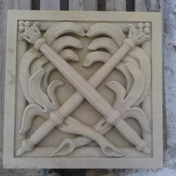 Kamieniarstwo Artystyczne - Kwadratowa płyta z piaskowca z płaskorzeźbą przedstawiającą skrzyżowane pochodnie z dekoracyjnymi elementami roślinnymi, umieszczona na betonowej powierzchni.