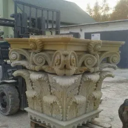 Dekoracyjny, rzeźbiony kapitel kolumny z piaskowca, umieszczony na palecie, z widocznym w tle wózkiem widłowym i budynkiem gospodarczym.