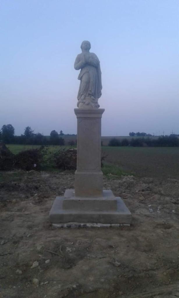 Kamienna figura świętej na postumencie, umieszczona na polu, widok na horyzont w pochmurny dzień.