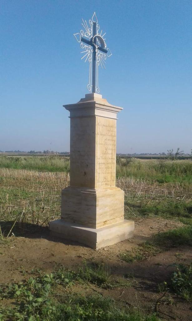 Kamienna kapliczka z krzyżem na szczycie, ustawiona na polu z widocznymi śladami po żniwach, inskrypcje wyryte na kamieniu.