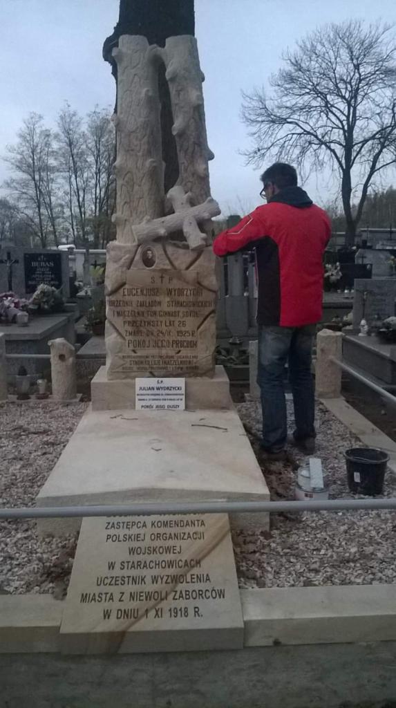 Pracownik w czerwonej kurtce czyści jasny, kamienny nagrobek z inskrypcjami na cmentarzu; w tle inne groby i drzewa bez liści.