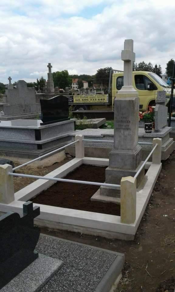 Nowy, jasny nagrobek z krzyżem i obramowaniem, świeżo obsypany ziemią, na tle innych nagrobków i żółtego samochodu dostawczego.