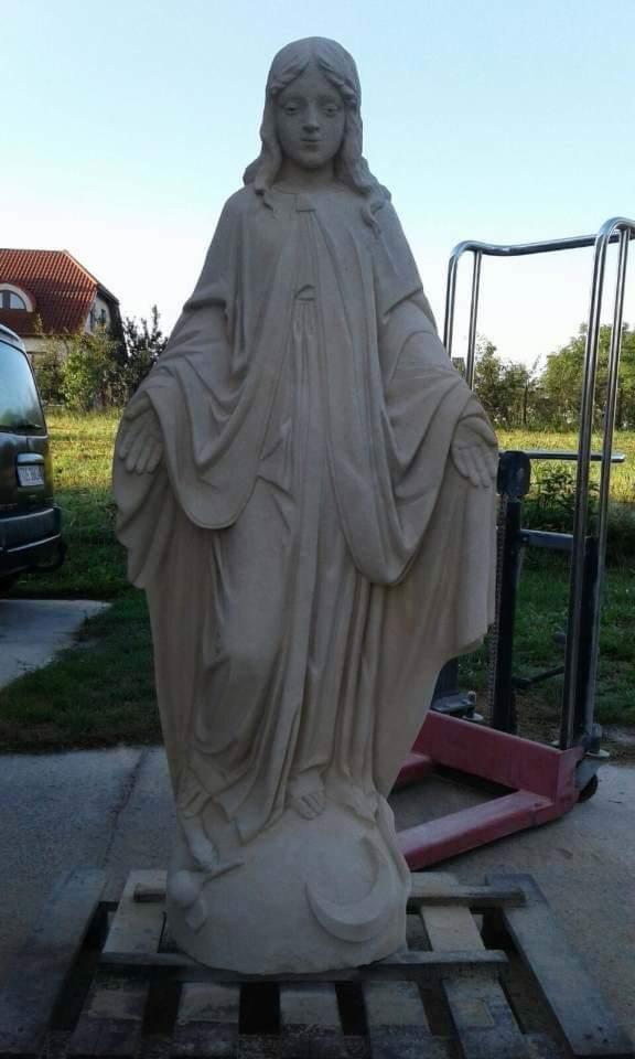 Kamienna figura Matki Boskiej z rozłożonymi rękami, umieszczona na palecie transportowej z widocznym w tle budynkiem mieszkalnym i wózkiem widłowym.