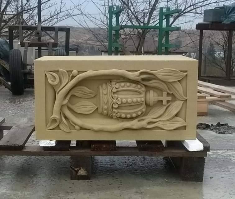 Piaskowy blok z rzeźbionym motywem korony otoczonej liśćmi, prawdopodobnie element nagrobka, umieszczony na drewnianej palecie w otoczeniu budowlanym.