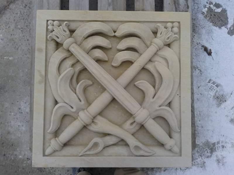 Kwadratowa płyta z piaskowca z płaskorzeźbą przedstawiającą skrzyżowane pochodnie z dekoracyjnymi elementami roślinnymi, umieszczona na betonowej powierzchni.