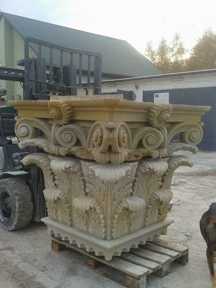 Dekoracyjny, rzeźbiony kapitel kolumny z piaskowca, umieszczony na palecie, z widocznym w tle wózkiem widłowym i budynkiem gospodarczym.