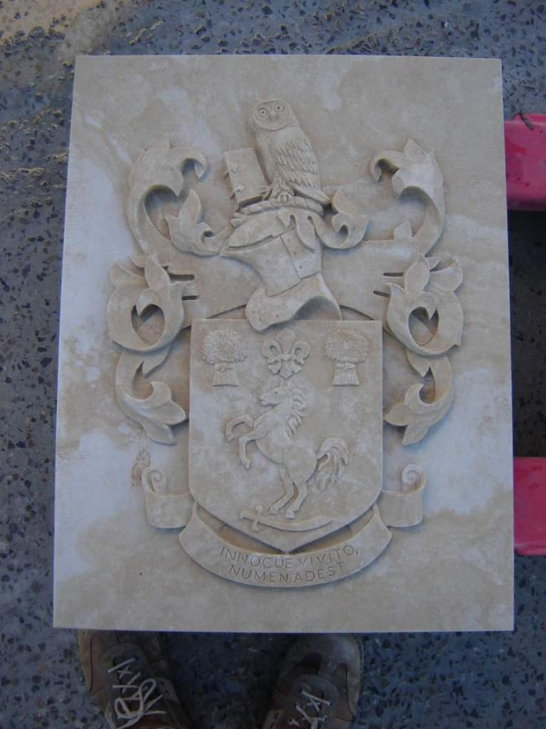 Wyryty w jasnym kamieniu herb rodowy z sową, hełmem rycerskim i koniem, częściowo zacieniony, trzymany przez osobę w brudnych butach.