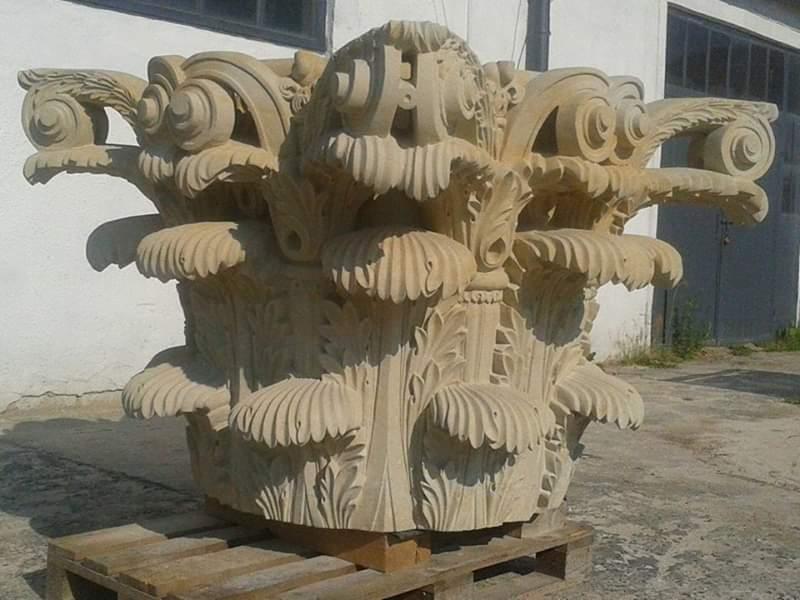 Gotowa, ozdobna głowica kolumny z piaskowca z detalami w stylu jońskim, umieszczona na drewnianej palecie przed budynkiem.