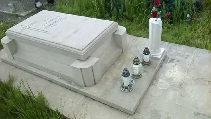 Jasny, nowoczesny pomnik z inskrypcją i krzyżem, uzupełniony o białą kolumnę z czerwonymi kwiatami oraz trzy szklane lampiony, umieszczony na płycie w otoczeniu zielonej trawy.