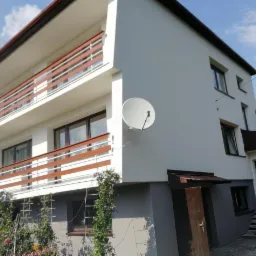 Dwupiętrowy dom po remoncie elewacji z balkonami z drewnianą balustradą, białą elewacją i szarą podmurówką, z anteną satelitarną na ścianie i pnącymi się roślinami przy elewacji.