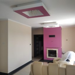 Mardosz - Nowoczesny salon z różową ścianą kominkową i beżowymi ścianami. Sufit podwieszany z różowym obramowaniem i kryształowym żyrandolem. W tle widoczne drzwi, na pierwszym planie beżowa kanapa.