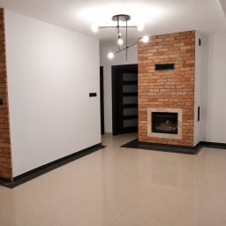 Mardosz - Nowocześnie wykończone wnętrze z fragmentami ścian z czerwonej cegły, beżowymi płytkami i wbudowanym kominkiem. Designerska lampa sufitowa i czarne listwy przypodłogowe.