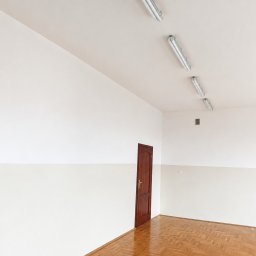 Mard-Bud - Puste pomieszczenie z białymi ścianami i beżowym pasem, drewnianą podłogą w jodełkę oraz brązowymi drzwiami. Na suficie cztery świetlówki, a na ścianie kratka wentylacyjna. Świeżo pomalowane ściany.