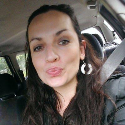 Portret kobiety z długimi, brązowymi włosami i kolczykami w kształcie kół, siedzącej w samochodzie, z zapiętym pasem bezpieczeństwa.