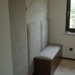 Wnęka z siedziskiem i tapicerowanymi panelami ściennymi w szarej kolorystyce, uzupełniona o drewnianą skrzynię pod siedziskiem, z oknem w tle.