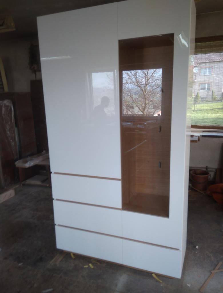 Biała szafa z szufladami i otwartą wnęką, widoczna w trakcie montażu, z odbiciem osoby w błyszczącej powierzchni.