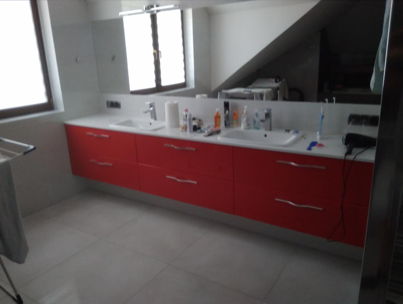 Czerwona szafka łazienkowa z dwiema umywalkami pod białą ścianą z lustrem i oknem, widok na poddasze.