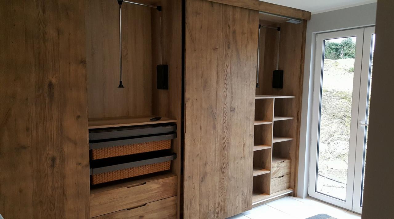 Drewniana szafa z przesuwnymi drzwiami, widoczne półki, szuflady i kosze z wikliny, system do wieszania ubrań, jasne wnętrze.