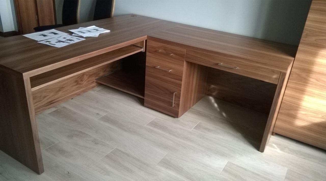 Kątowy, drewniany biurko z szufladami i półkami, na blacie leżą białe kartki z rysunkami technicznymi.