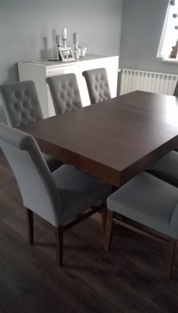 Stół z ciemnego drewna z szarymi krzesłami z guzikami, biała komoda w tle z dekoracjami, wnętrze w szarościach.