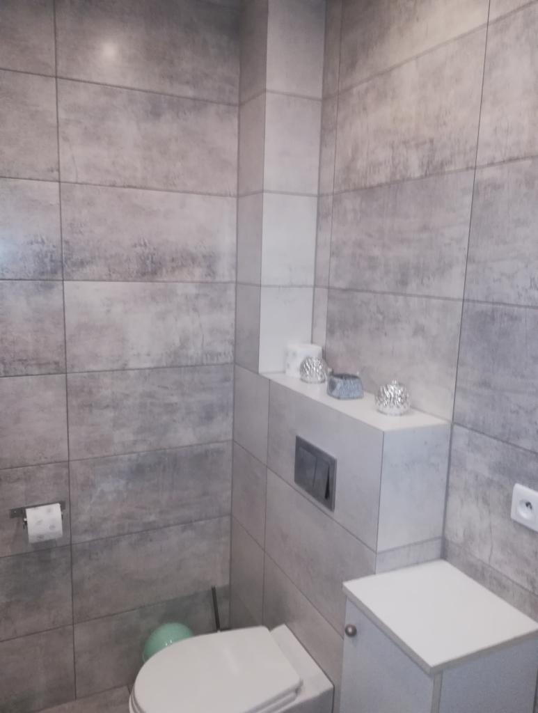 Wykończona łazienka z szarymi, imitującymi beton, płytkami ceramicznymi na ścianach, podwieszaną toaletą i zabudową z półką na dekoracje.