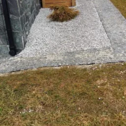 Ścieżka z kostki granitowej obrzeżona grysem, przylegająca do ściany budynku wykończonej kamieniem naturalnym z drewnianą osłoną i ozdobnym iglakiem.