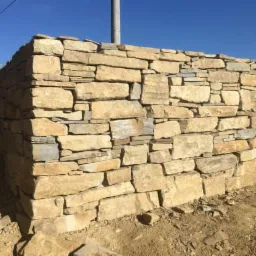 Fragment muru z nieregularnych, jasnych kamieni na fundamencie, widoczny na tle ziemi i błękitnego nieba.