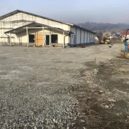 Budynek magazynowy w trakcie budowy z rusztowaniem, przygotowany teren z gruzem, mała koparka i walec w tle.