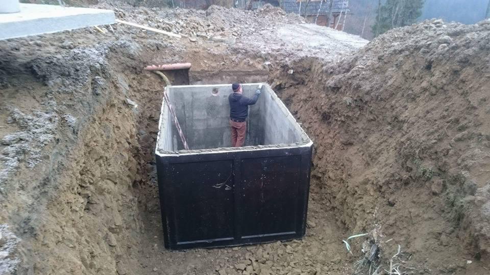 Czarny, prefabrykowany zbiornik betonowy częściowo zagłębiony w wykopie ziemnym, z pracownikiem wewnątrz uszczelniającym połączenia, widoczne wystające rury i świeży grunt.