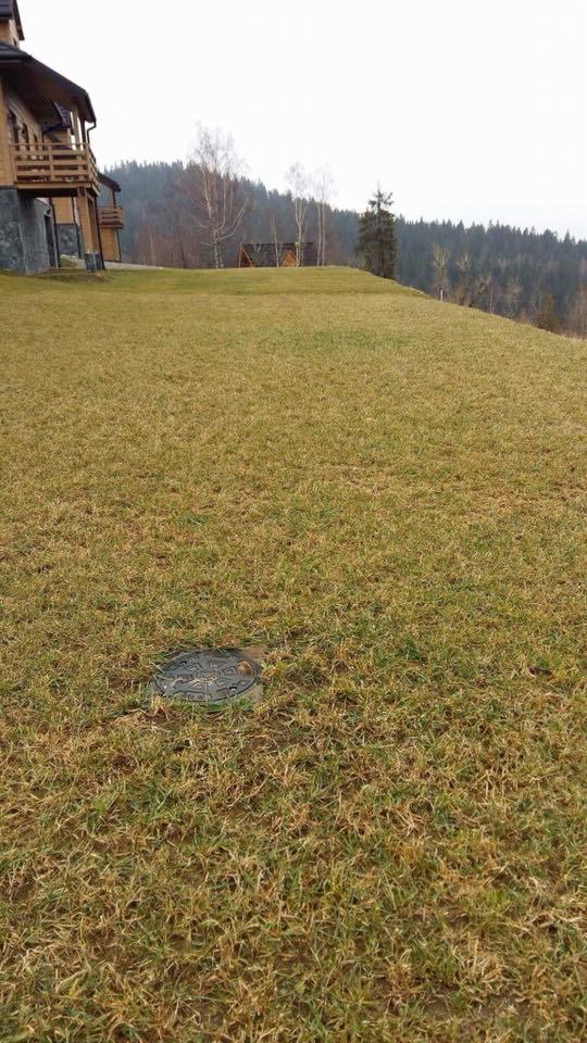 Żeliwna pokrywa studzienki rewizyjnej w trawniku na nierównym terenie, w tle dom z bali i las.