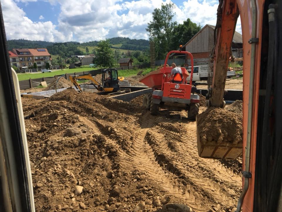 Wykonanie wykopu pod fundamenty domu jednorodzinnego z użyciem koparki i wywrotki, widok z kabiny innej maszyny, w tle zabudowa mieszkalna i zieleń.