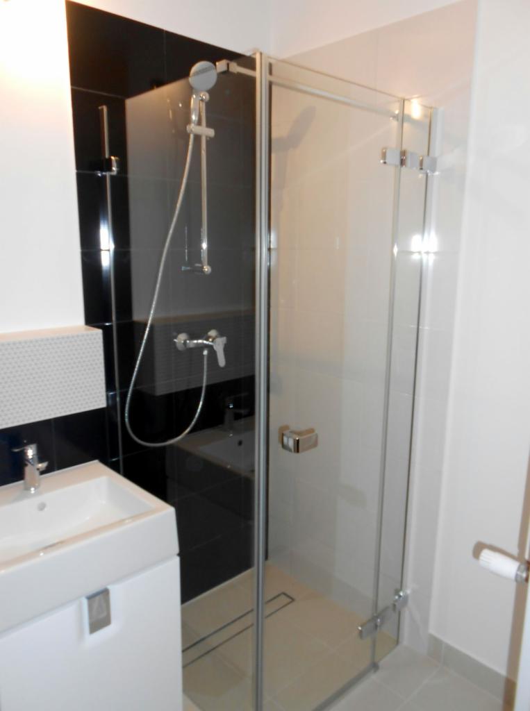 Nowoczesna kabina prysznicowa ze szklanymi drzwiami i chromowanymi zawiasami, obok umywalki na szafce z białym frontem, czarno-białe płytki na ścianie w tle.