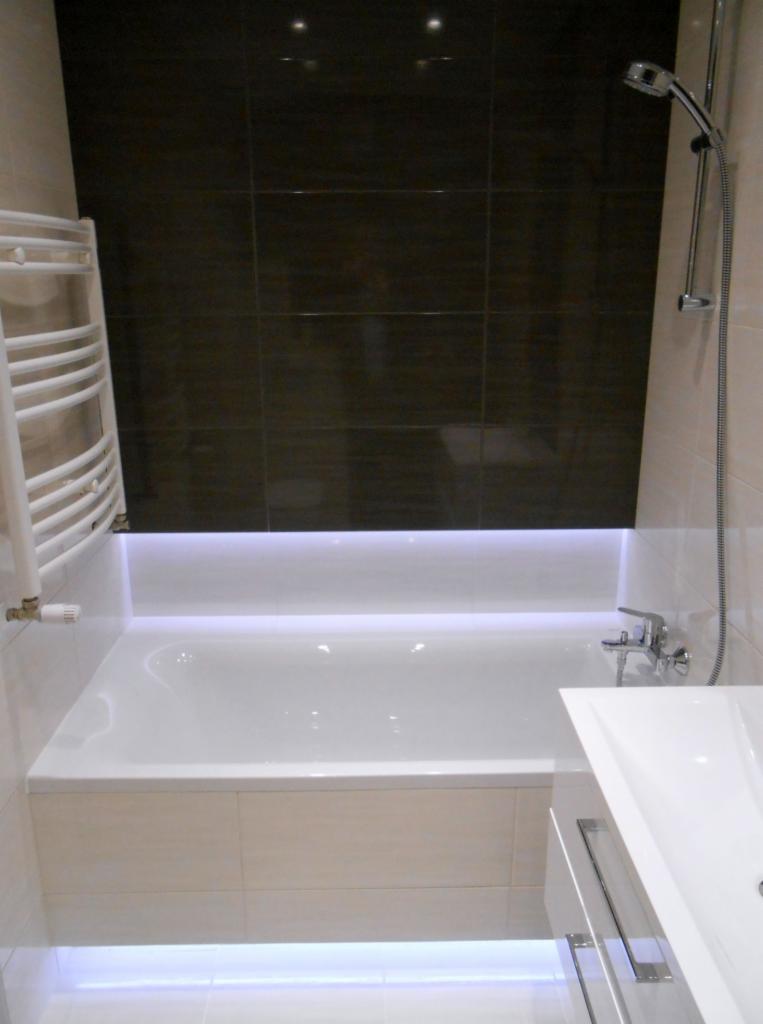 Biała wanna z podświetleniem LED, obok umywalka z szafką, ciemne płytki na ścianie, grzejnik drabinkowy i chromowana słuchawka prysznicowa w wykończonej łazience.